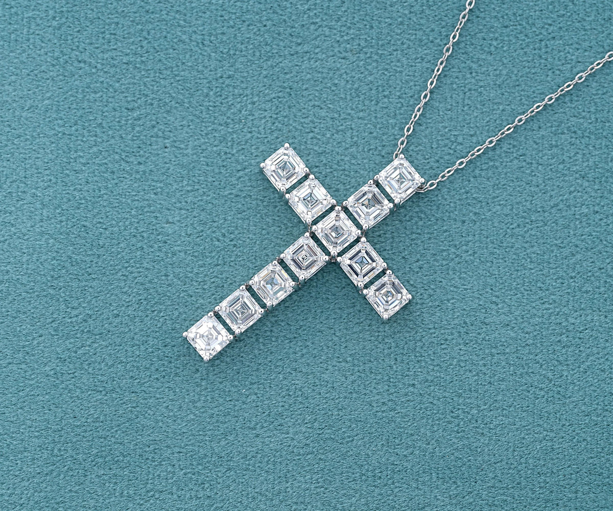 A silver cross pendant necklace paved with square-cut clear gemstones, displayed on a textured teal-blue background.