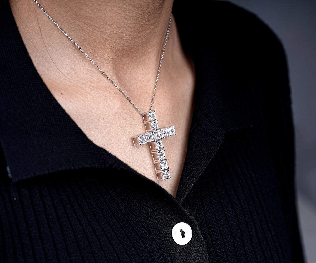 A silver cross pendant necklace set with small clear stones, worn on a person's neck over a black ribbed polo shirt.
