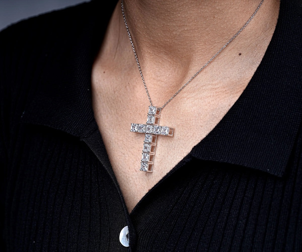 A close-up of a silver cross pendant necklace with clear stones, worn on a person's neck over a black ribbed polo shirt.