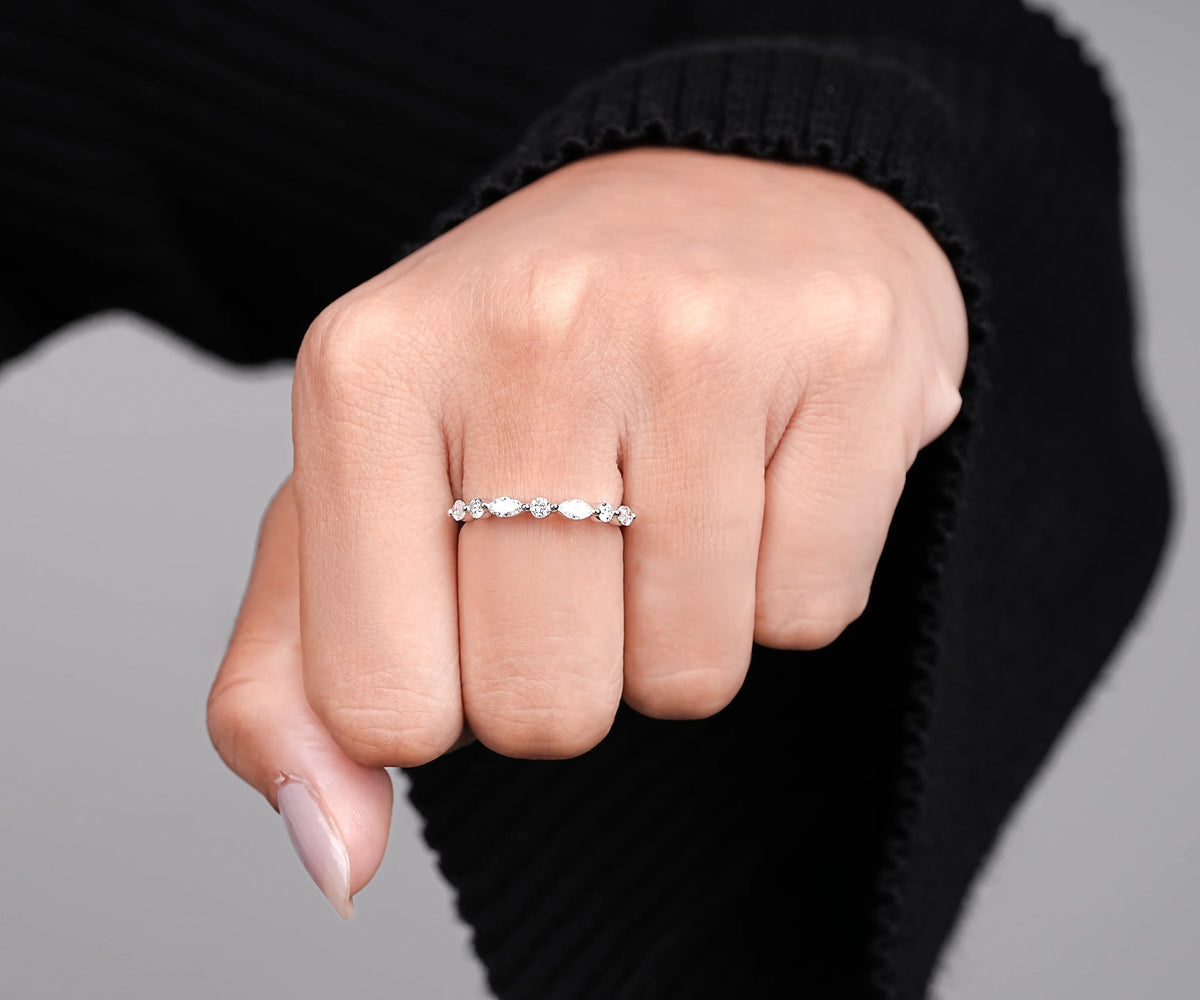 A close-up of a hand wearing a delicate silver band ring with a row of small clear marquise-cut gemstones, set against a black ribbed sweater and a neutral gray background.