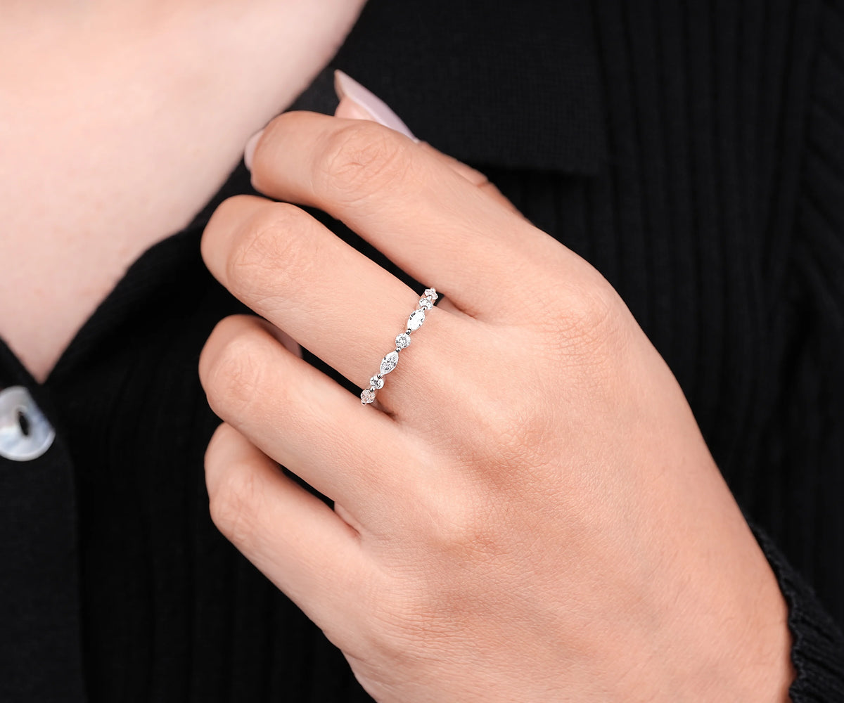 A close-up of a person's hand wearing a delicate silver band ring with small sparkling stones, set against a black ribbed sweater.