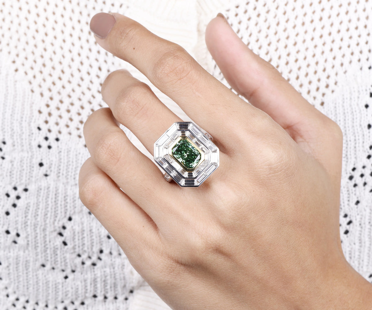 A silver-toned geometric ring with a radiant-cut green gemstone, worn on a person's hand against a white textured knit background.