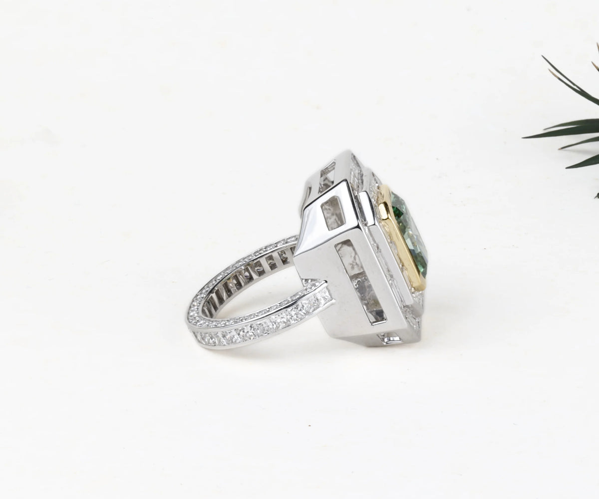 A silver-toned ring with a large rectangular green gemstone and a yellow-gold accent on the side, set against a plain white background with a small green leaf in the corner.