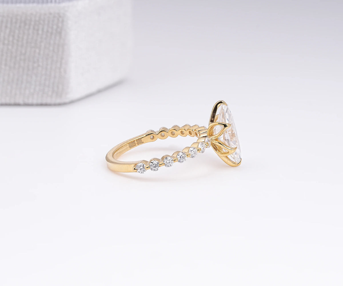 A gold ring with a large clear oval-cut center stone and a row of small clear stones along the band, photographed on a clean white surface with a soft gray textured box in the background.
