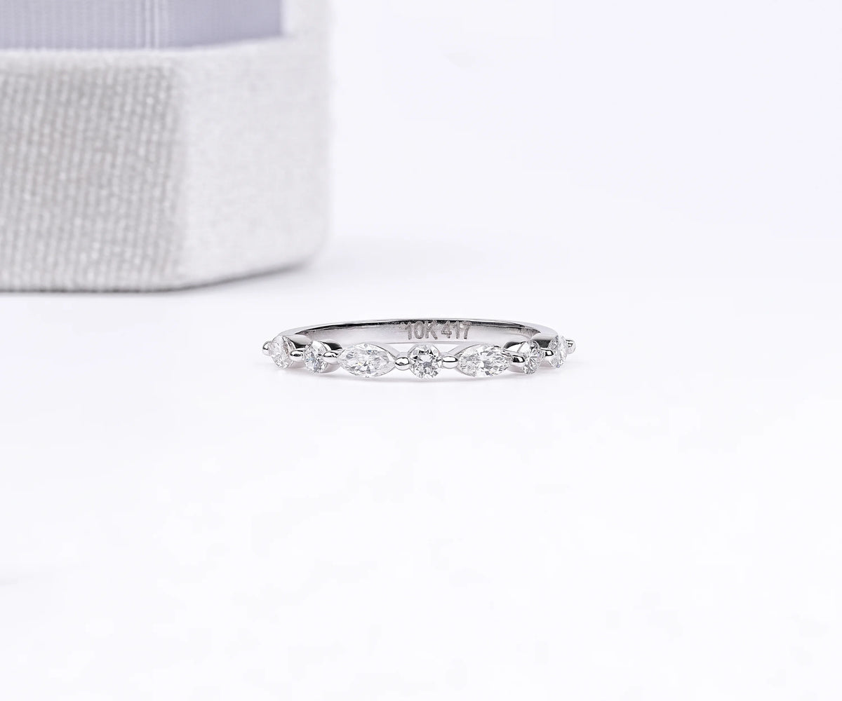 A delicate silver band ring with a row of small marquise-cut clear stones, photographed on a white surface with a soft-focus textured box in the background.