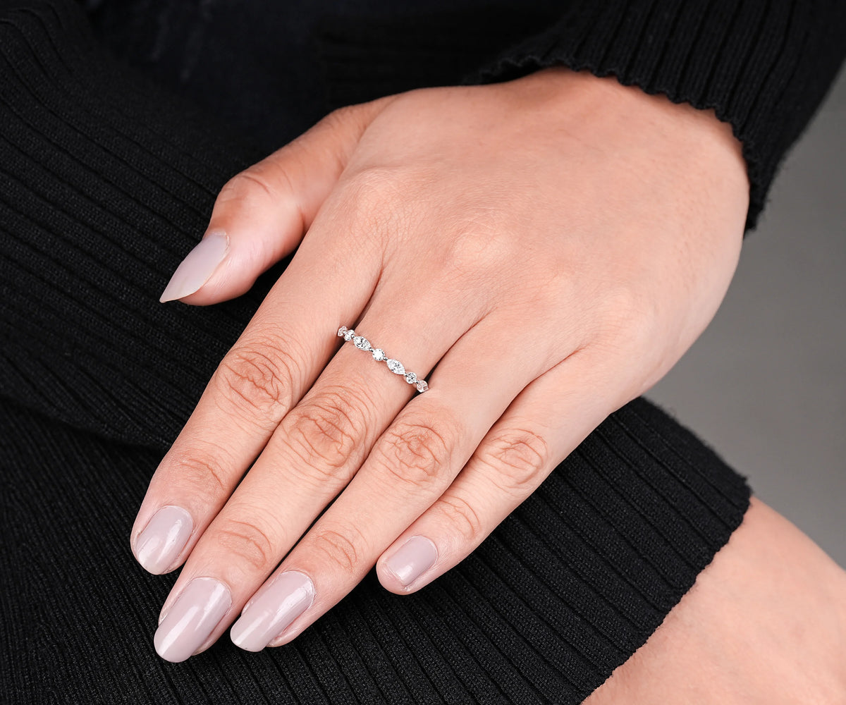 A close-up of a hand wearing a delicate silver band ring set with small clear stones, resting on a black ribbed sweater against a neutral gray background.