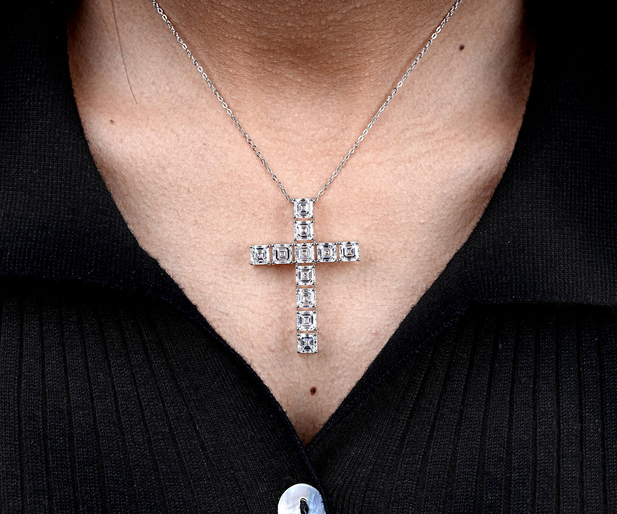 A silver cross pendant necklace set with clear stones, worn on a person's neck over a black ribbed shirt.