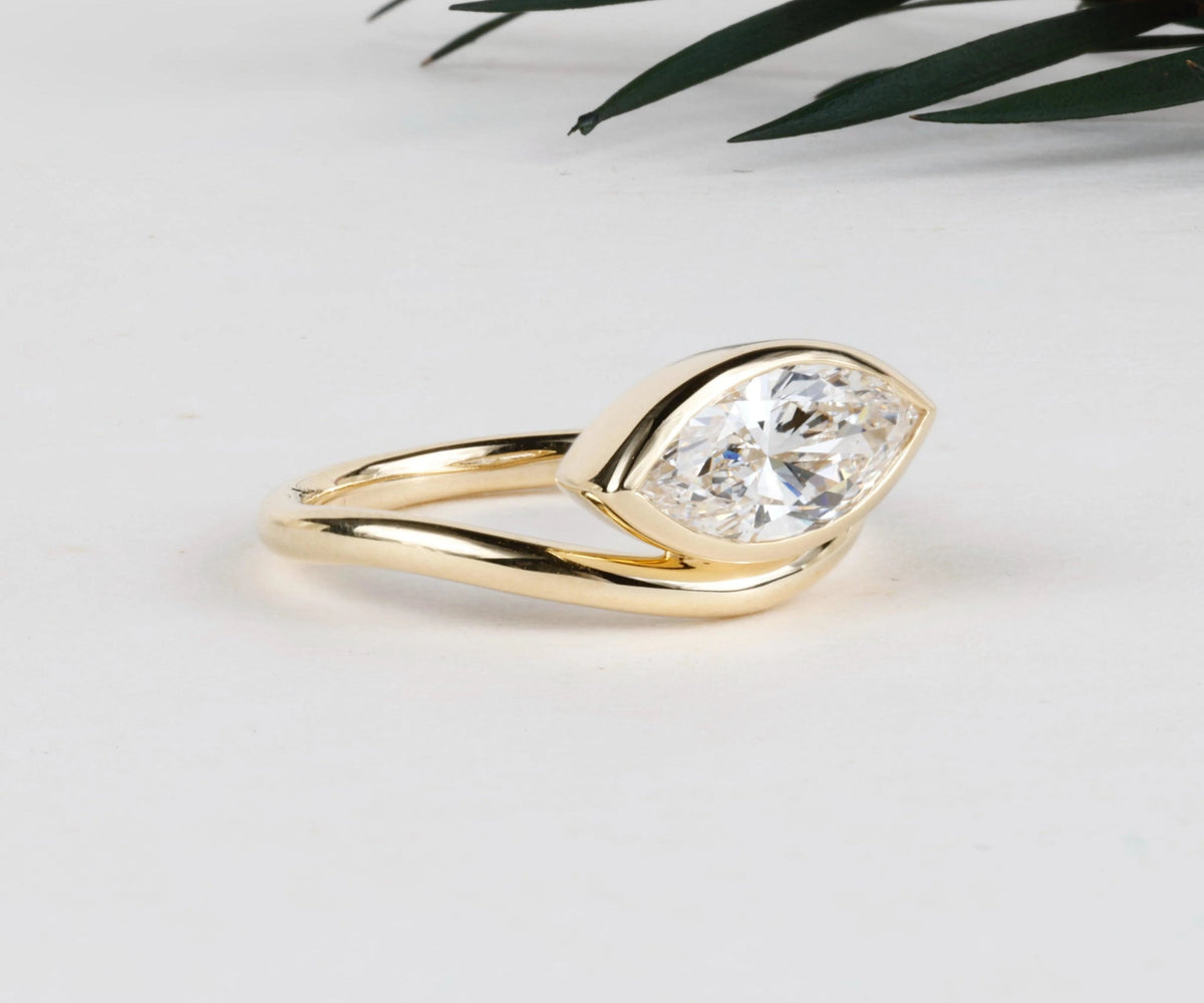 A gold ring featuring a marquise-cut clear gemstone in a prong setting, displayed on a clean white surface with a touch of green foliage in the background.