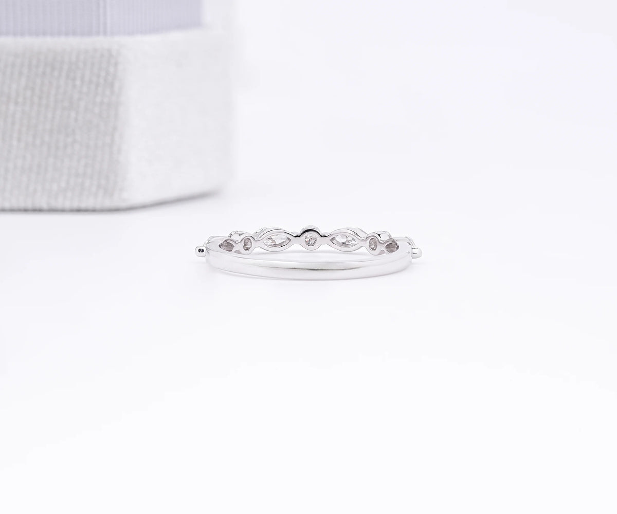 A delicate silver band ring with a repeating small oval link motif, displayed on a clean white surface with a soft-focus textured box in the background.
