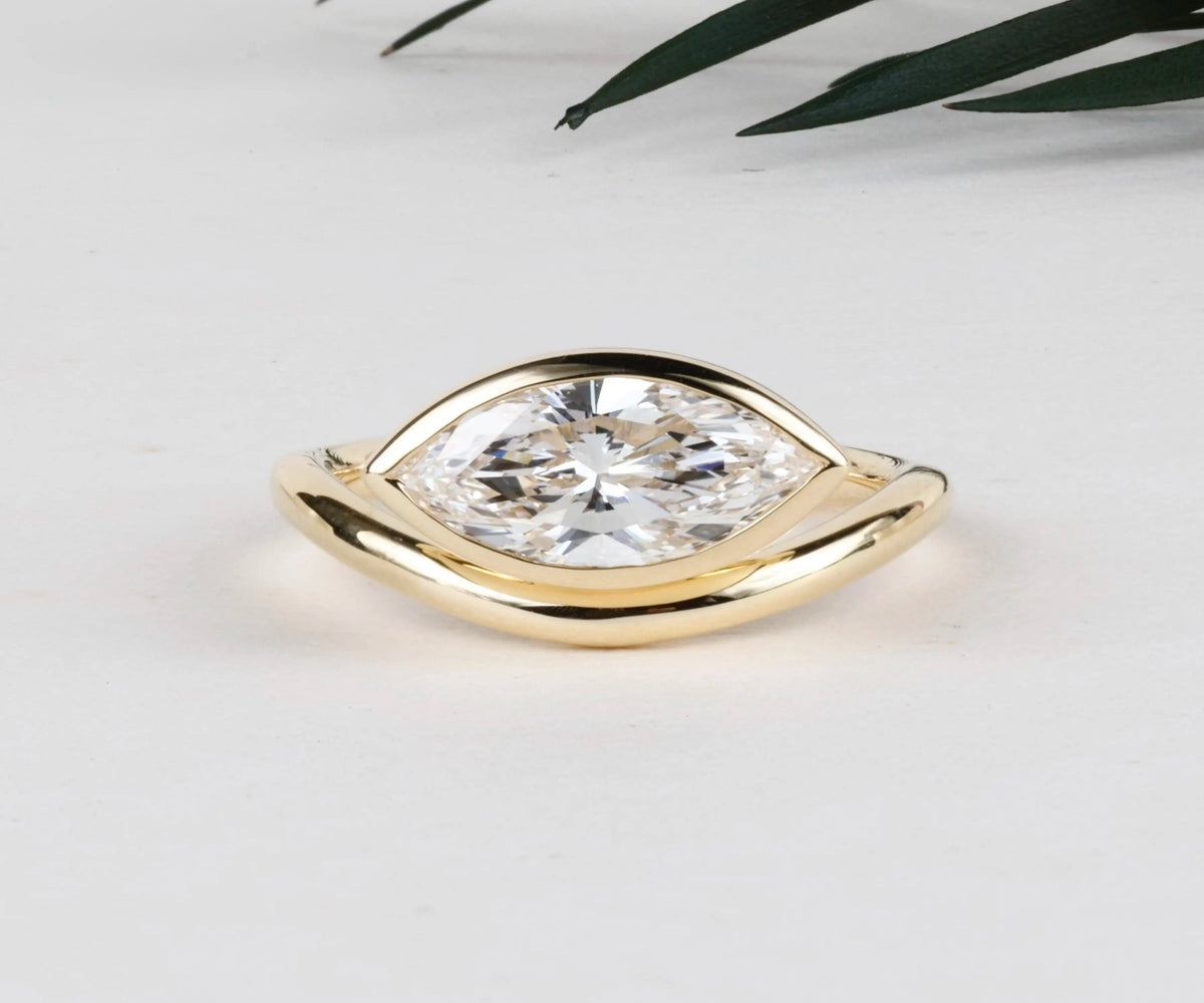 A gold ring featuring a large marquise-cut clear gemstone in a split setting, photographed on a light neutral background with a small dark green leaf in the upper right corner.