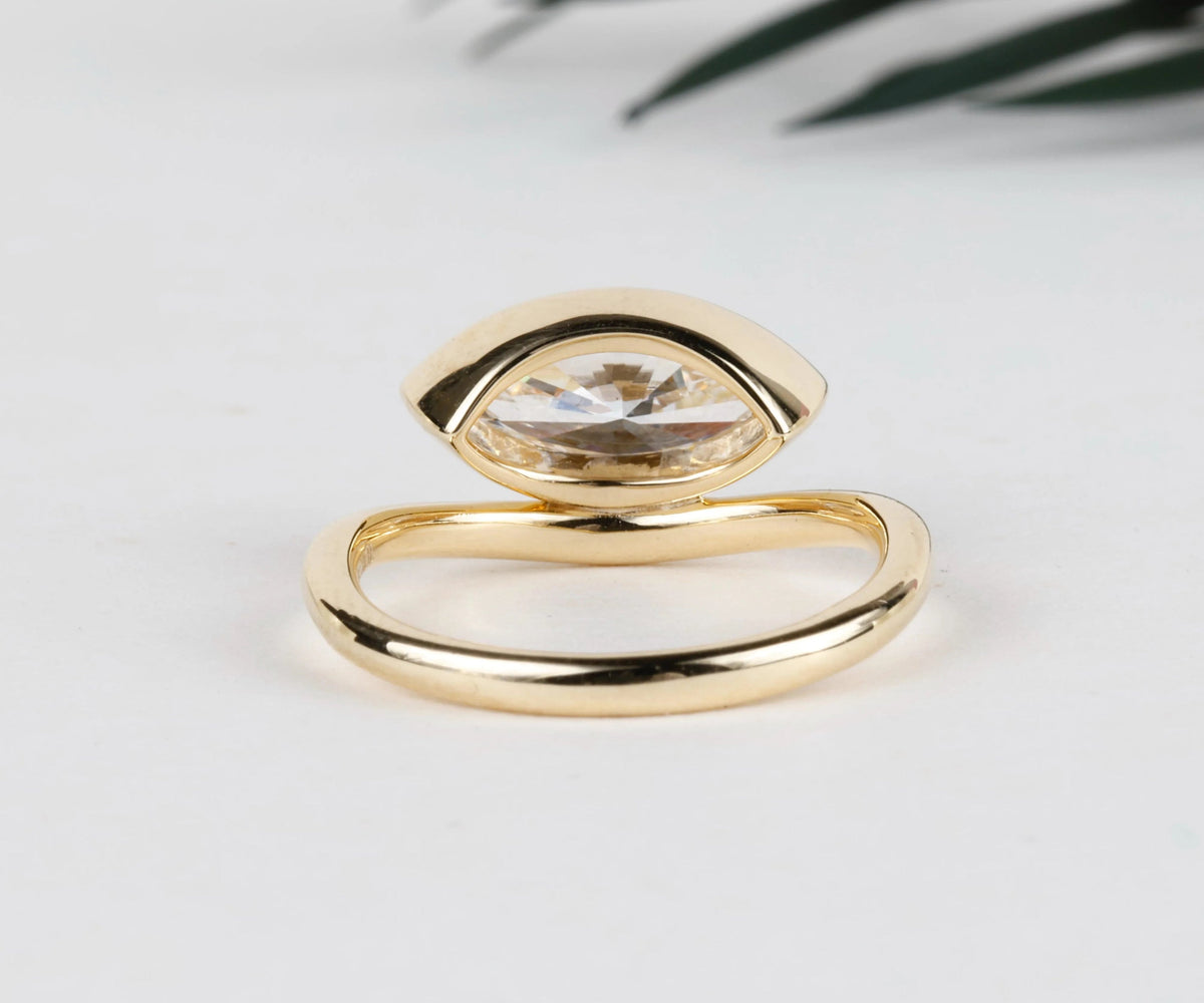 A gold-toned open ring with a marquise-cut clear gemstone, photographed on a clean white surface with a soft-focus green leaf in the background.