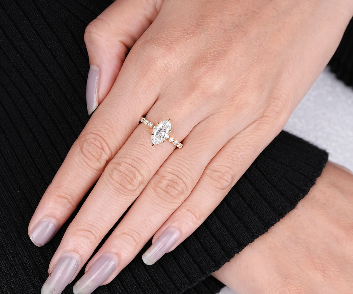 A close-up of a hand wearing a delicate gold ring with a marquise-cut clear gemstone and small side stones, resting against a black ribbed sweater.