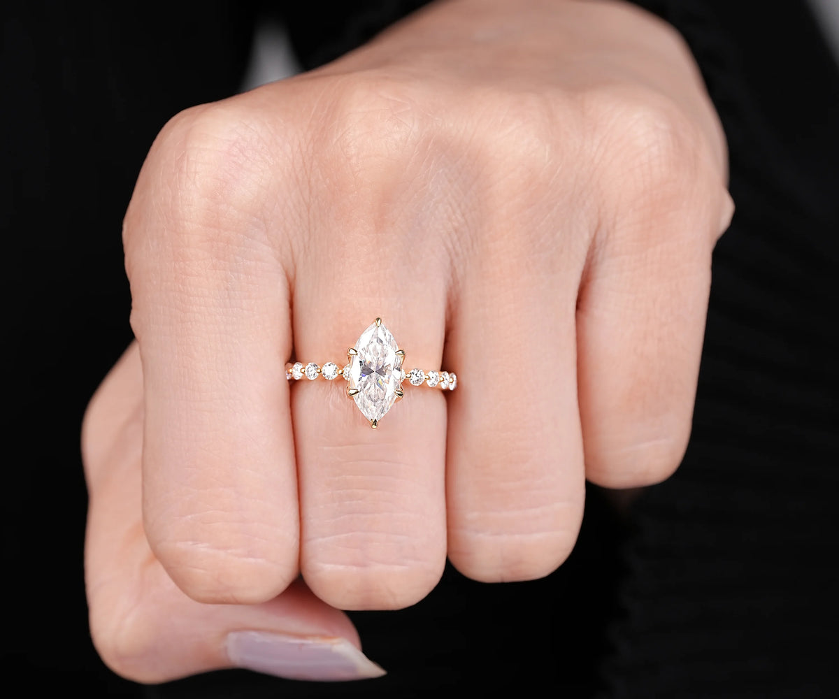 A close-up of a hand wearing a gold ring with a marquise-cut clear center stone and small side stones, set against a dark background.