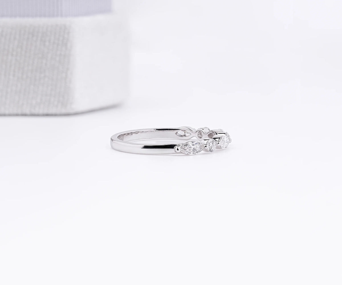 A slim silver band ring with a row of small clear round-cut stones, photographed on a clean white surface with a soft-focus white textured box in the background.