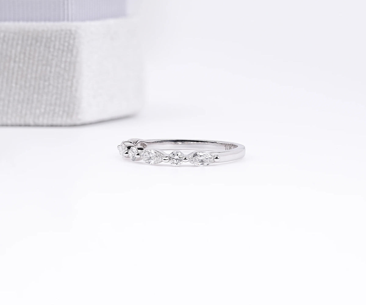 A delicate silver band ring with a row of small clear stones, photographed on a clean white surface with a soft-focus textured box in the background.